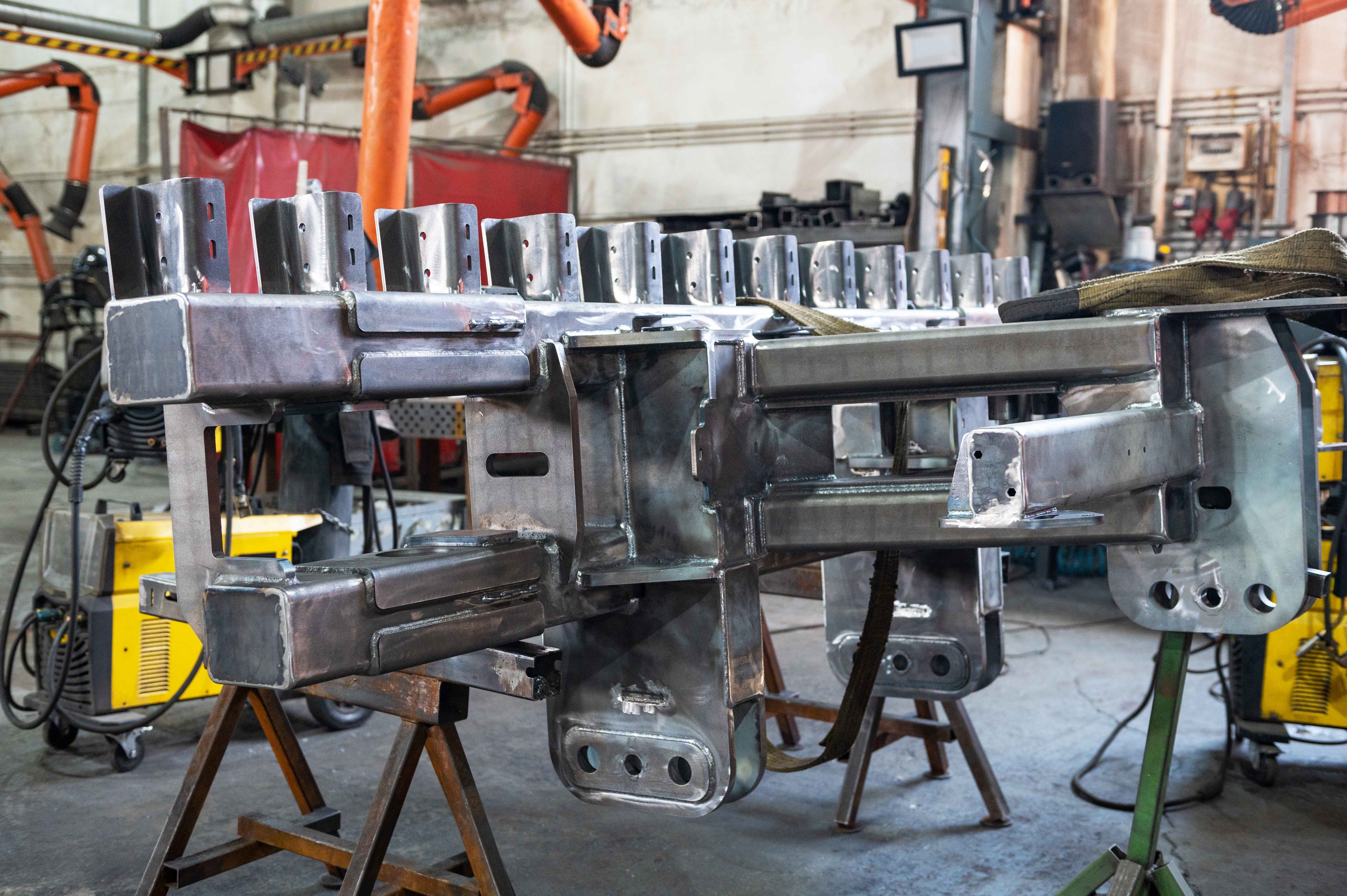 Large fabricated steel assembly mounted on support stands inside a manufacturing facility, surrounded by welding equipment and production machinery.